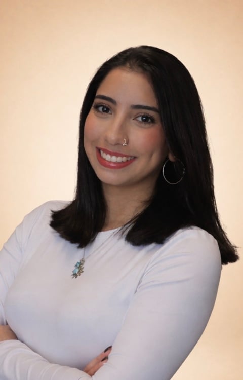 Portrait of a smiling woman with long black hair, wearing a white long-sleeve top and hoop earrings against a beige background