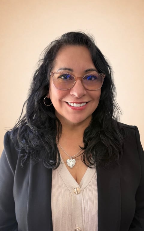 Portrait of a woman with long dark wavy hair, wearing glasses, a black blazer, and silver necklaces, smiling at the camera