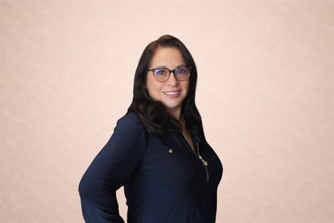 Portrait of a woman with long dark hair wearing glasses and a dark blue button-up shirt against a beige background