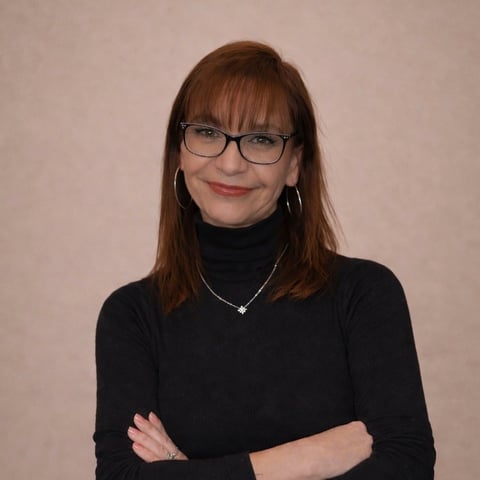 Woman with reddish-brown hair and bangs wearing glasses, black turtleneck, and silver necklace, smiling at camera with arms crossed