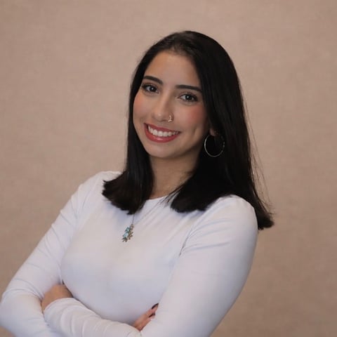 Portrait of a smiling woman with long dark hair wearing a white long-sleeve top and hoop earrings against a beige background