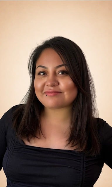 Portrait of a woman with long dark hair and pink lipstick wearing a black off-shoulder top against a beige background