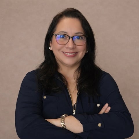 Woman with long dark hair wearing blue-framed glasses and navy blazer, smiling at camera with arms crossed
