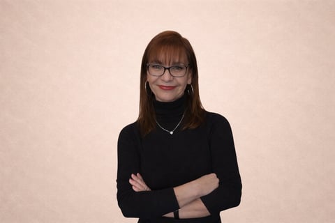 Woman with brown hair and glasses wearing a black long-sleeve shirt, arms crossed, smiling at camera against a beige background
