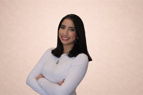 Smiling woman with long dark hair wearing a white long-sleeved top and hoop earrings, standing with arms crossed against a beige background